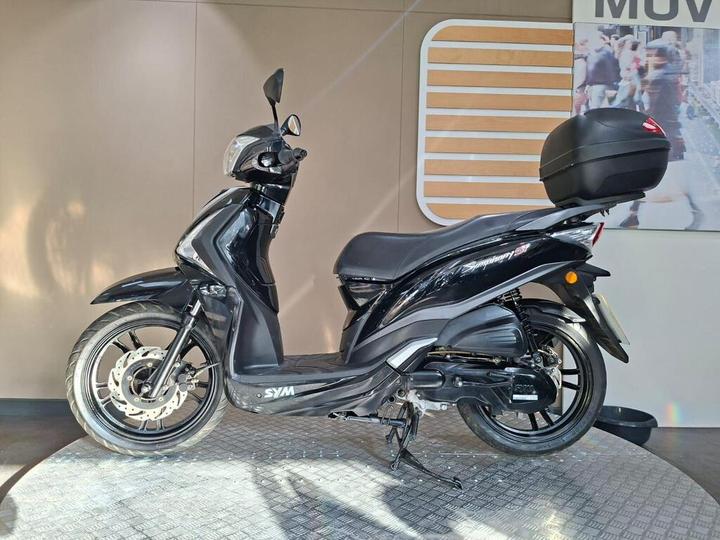 A black scooter parked on a textured surface, featuring a top case for storage and modern design elements, against a plain background with soft lighting.
