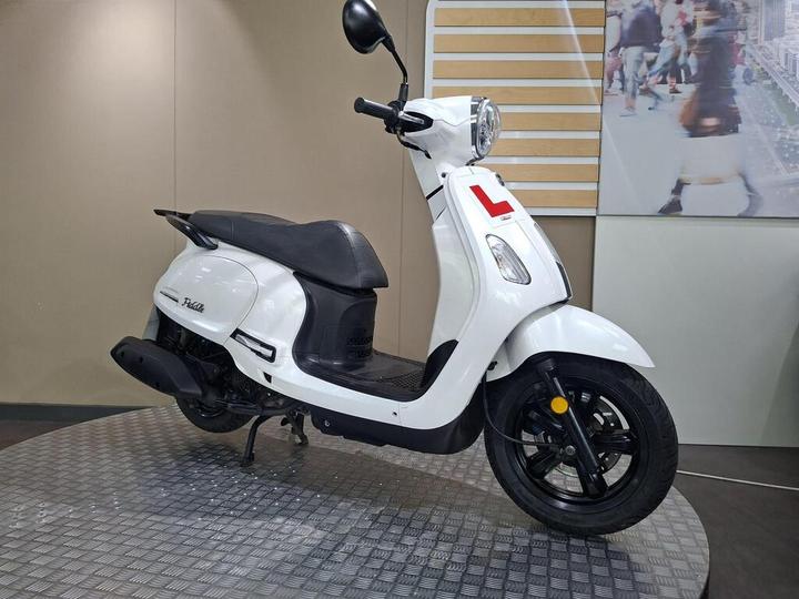 A white scooter with a black seat, featuring an "L" sign on the front, displayed on a textured surface with a blurred background of people and buildings.