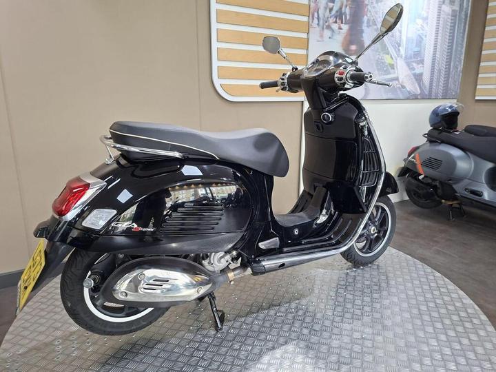 A black scooter with a sleek design is displayed on a metal platform in an indoor showroom, with a blurred background featuring another scooter.