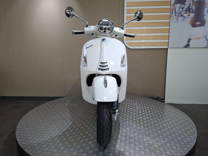 A front view of a white Vespa scooter displayed on a circular platform with a textured surface and a neutral background.