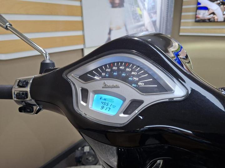 Close-up of a scooter's dashboard displaying a speedometer and digital readout with speed at 45.5 km/h and fuel level indicator. The handlebar and control switches are visible, against a blurred background.