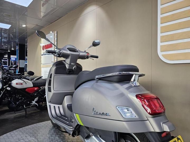 A gray Vespa scooter is parked in a dealership, showcasing its sleek design, with other motorcycles in the background and a modern interior setting.