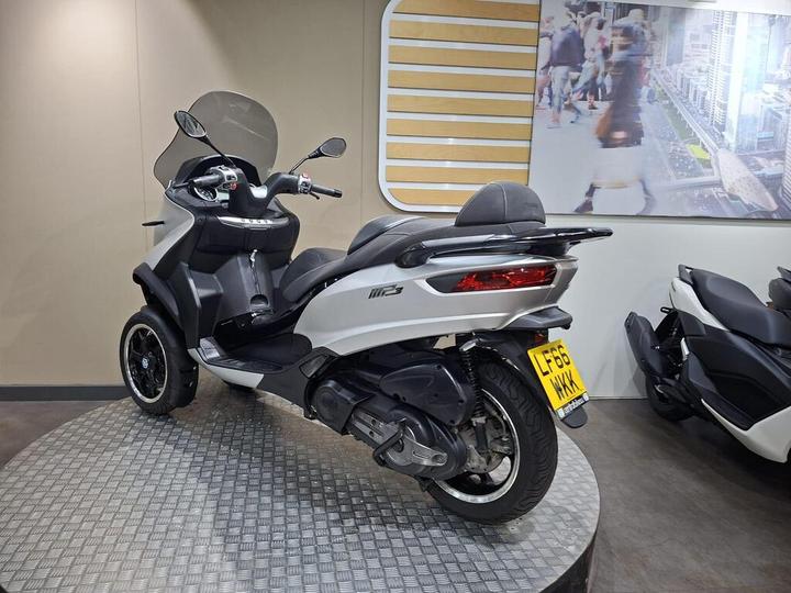 A white three-wheeled scooter displayed on a circular platform in a showroom, with a clear windshield and black seating. The background features a large image of a busy city street.