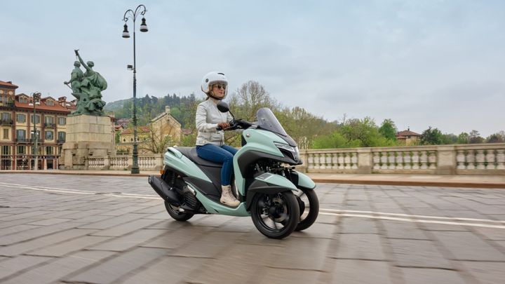 A woman wearing a helmet and sunglasses is riding a light blue scooter on a cobblestone street, with historic buildings and a statue visible in the background.