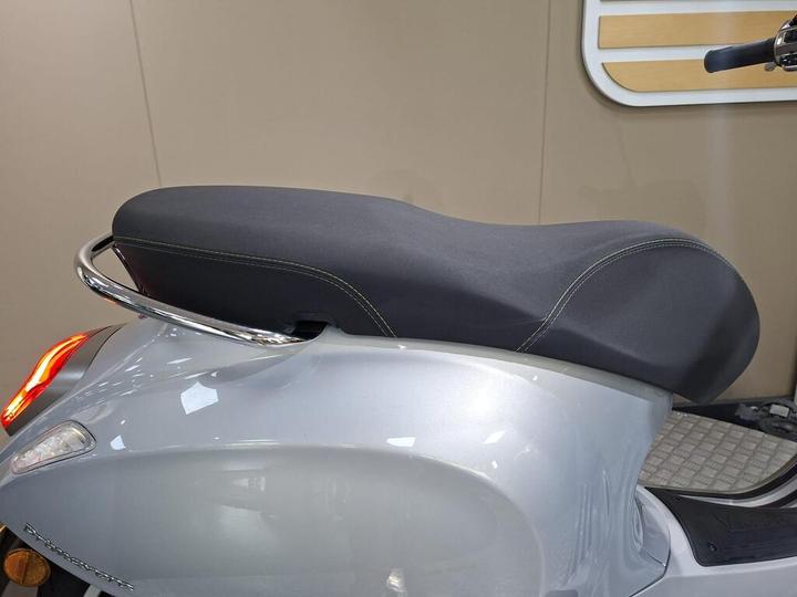 A close-up view of a sleek silver motorcycle seat with a black cover, featuring contrasting green stitching and a chrome rear grab rail. The background is a neutral beige.