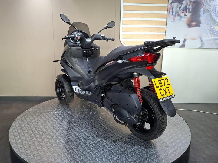 A sleek black three-wheeled scooter displayed on a circular platform, showcasing its rear view with red suspension and a license plate visible.