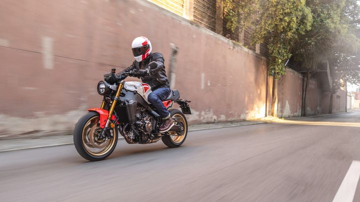 A motorcyclist in a leather jacket and white helmet rides a stylish motorcycle with red and black accents, speeding down a city street with a blurred background.