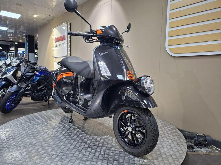 A black Vespa scooter with orange accents is displayed on a circular platform in a showroom, with additional motorcycles visible in the background.