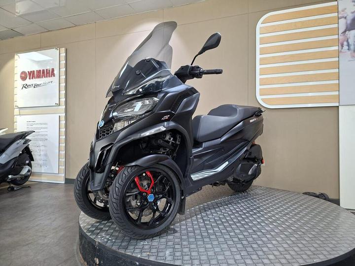 A sleek black three-wheeled scooter showcased on a circular platform in a dealership setting, with a Yamaha logo visible on the wall behind.