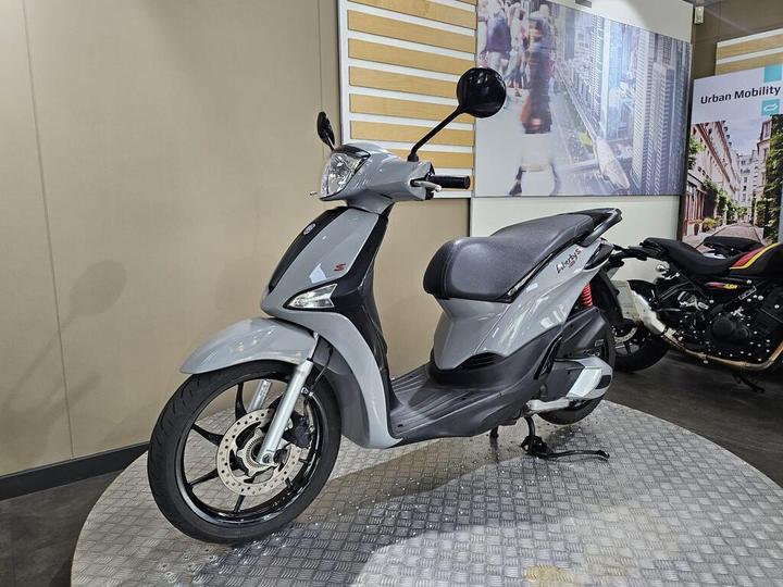 A gray scooter is displayed on a circular platform inside a showroom, with a modern background featuring urban imagery. Another motorcycle is partially visible in the background.