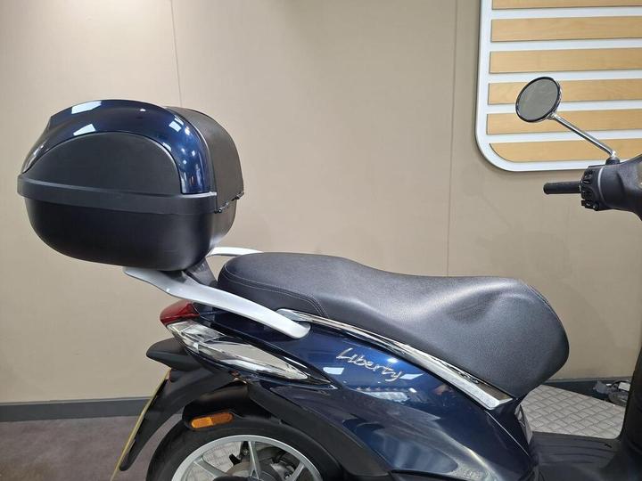 A close-up view of a navy blue scooter with a black top case, featuring a textured seat and chrome detailing on the side, set against a neutral background.