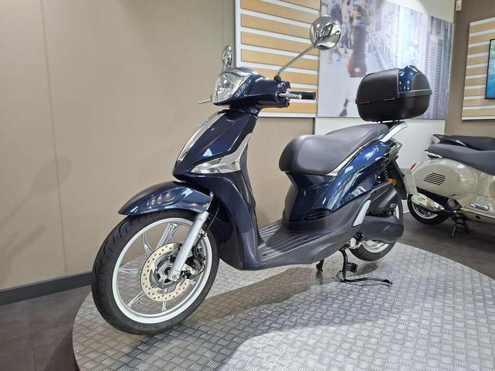 A blue scooter is displayed on a circular metal platform in a showroom, showcasing its sleek design, front fender, and rear storage box.