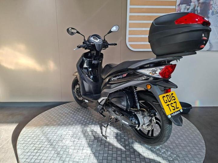 A black scooter with a top case, on a circular metallic platform, displayed against a neutral background.