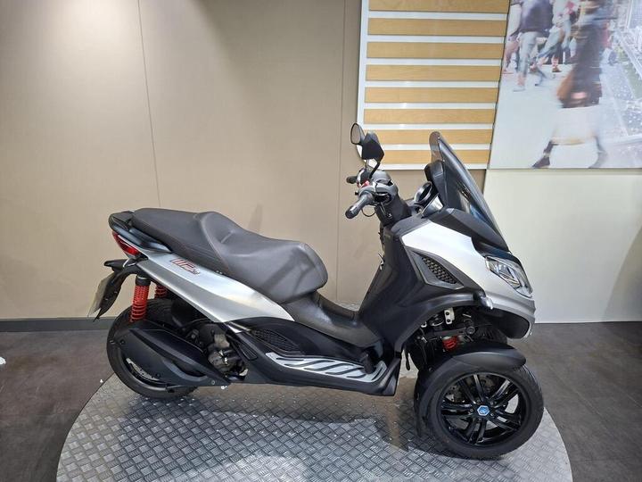 A silver scooter with three wheels displayed on a textured round platform against a neutral background, showcasing its sleek design and red shock absorbers.