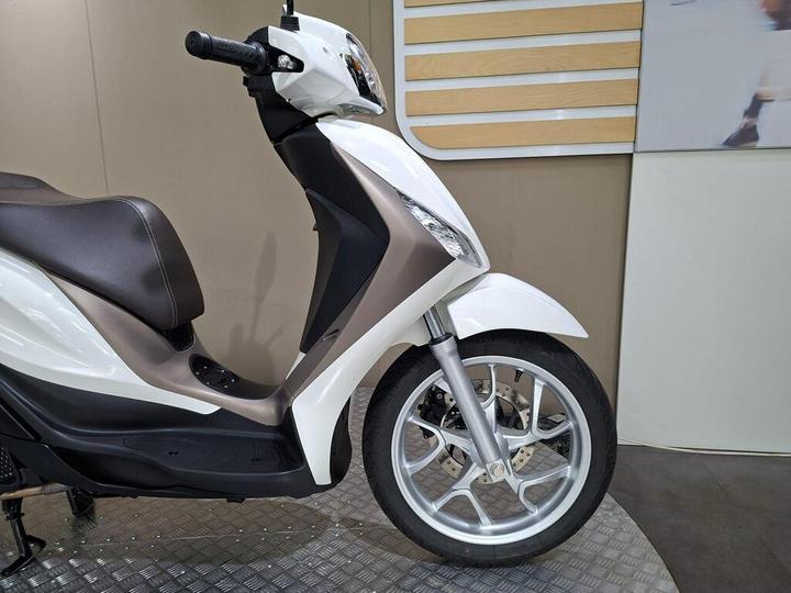 A sleek, modern scooter in white and light brown, featuring a comfortable seat, a curved body design, and a stylish front wheel with multiple spokes, displayed against a simple background.