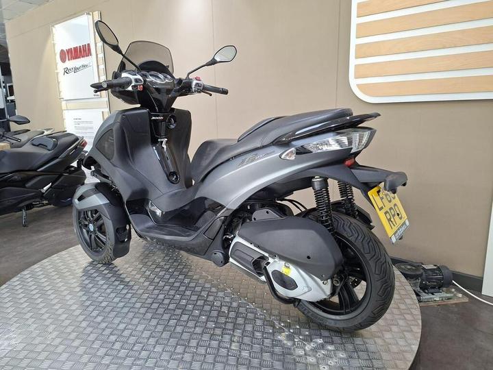A grey three-wheeled scooter displayed on a textured platform in a Yamaha showroom, with a partial view of another scooter in the background and signage on the wall.