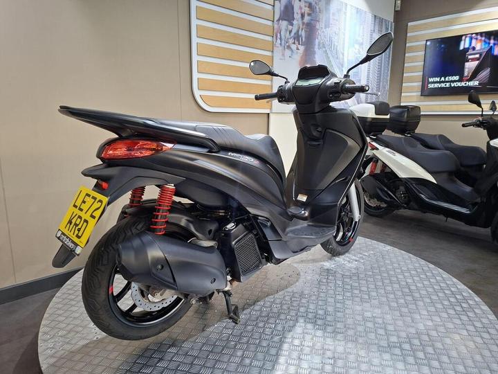 A sleek black scooter with a prominent rear design, featuring red shock absorbers and a shiny wheel. The scooter is displayed on a circular metallic platform in a modern showroom, with a blurred white scooter in the background and a promotional screen on the wall. The license plate reads "LE72 KRD."