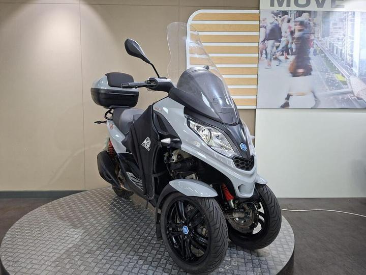 A gray three-wheeled scooter is displayed on a circular platform with a textured surface; it features a windshield, a storage compartment on the back, and is positioned against a backdrop of urban imagery and the word "MOVE."