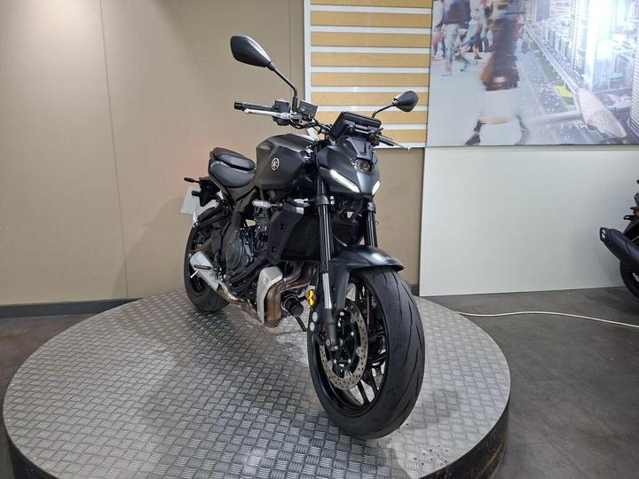 A sleek, modern motorcycle is displayed on a circular metal platform in a showroom setting, featuring a matte black finish, prominent headlight design, and a sporty stance.