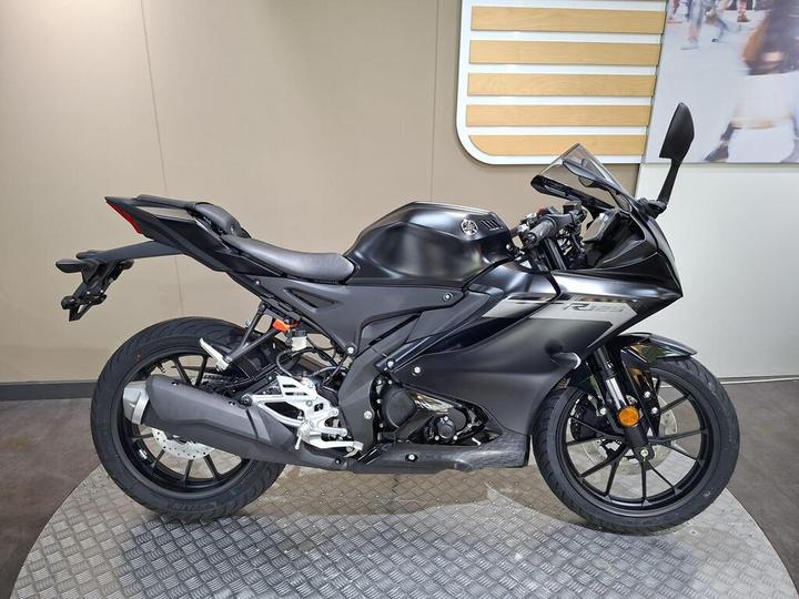 A sleek black Yamaha R15 motorcycle displayed on a textured platform, featuring a modern design with sporty lines and advanced components.