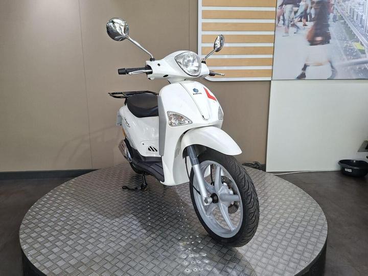 A white scooter displayed on a circular metallic platform with a textured surface, set against a neutral background featuring a wall display.