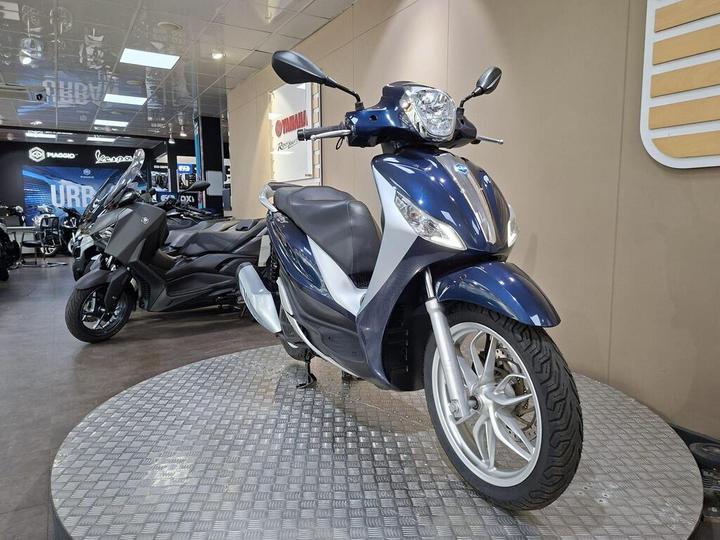 A blue scooter on a circular display platform in a motorcycle showroom, with a black scooter partially visible in the background.