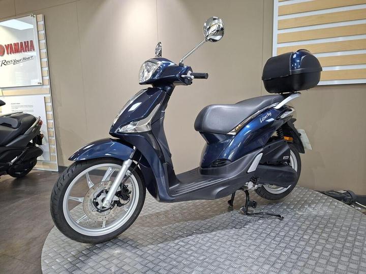 A blue Yamaha scooter displayed on a circular platform, featuring a stylish design, a comfortable seat, a rear storage box, and shiny alloy wheels, set against a neutral background with Yamaha branding visible.