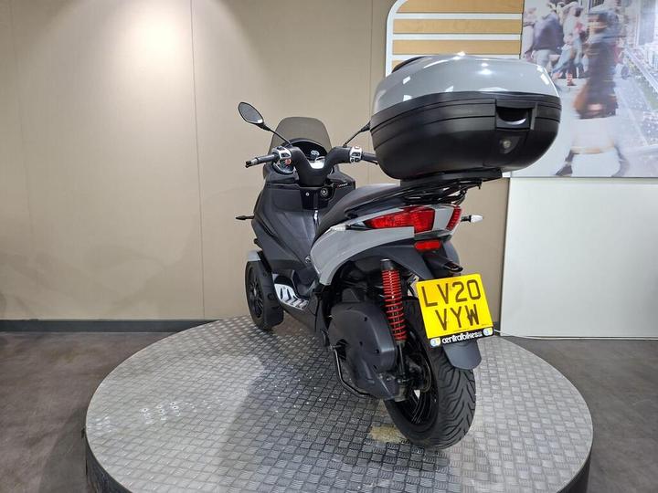 A rear view of a grey scooter with a top case, displayed on a circular platform in a modern showroom setting. The scooter features red shock absorbers and a visible license plate reading "LV20 VYW."
