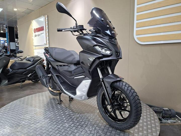 A sleek black scooter with a modern design is on display in a showroom, featuring a tall windscreen, large tires, and contrasting silver accents. In the background, another scooter is partially visible.