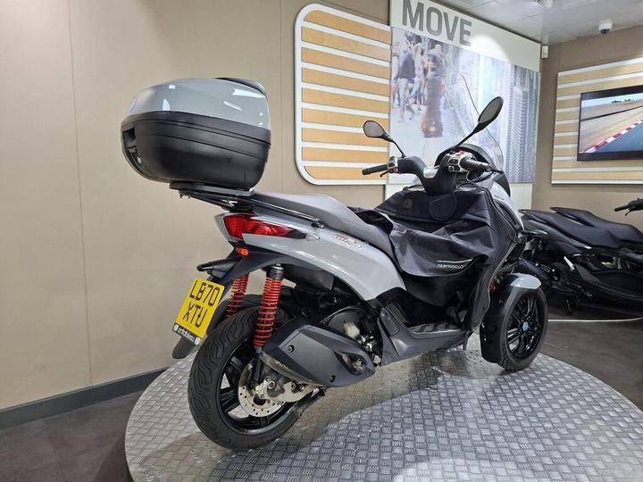 A grey scooter with a top case, positioned in a showroom with a metal floor and displayed alongside other scooters.