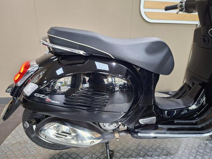 A close-up view of the side of a black scooter with a sleek design, featuring a comfortable seat and shiny chrome accents. The background shows a light-colored wall, emphasizing the scooter's glossy finish.