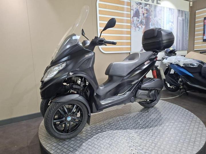 A black three-wheeled scooter with a windshield and a rear storage case, placed on a circular display in a showroom, with a blurred background of other vehicles and promotional signage.