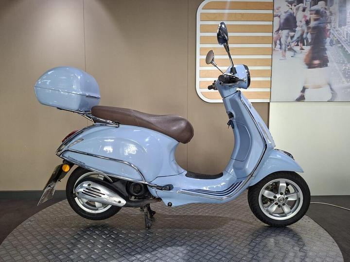 Vespa Primavera 50 2017 A blue scooter with a brown seat and a matching front storage compartment, displayed on a circular platform against a neutral background.