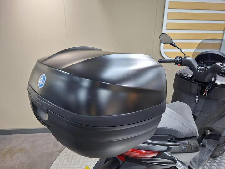 A close-up view of a matte black top case mounted on the rear of a scooter, with a blue logo visible on the case and the scooter's grey seat and controls partially shown.