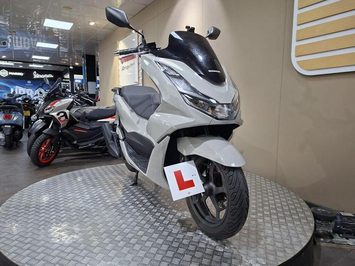 A light grey Honda scooter with a learner's plate stands on a circular display in a motorcycle showroom, surrounded by other motorcycles.