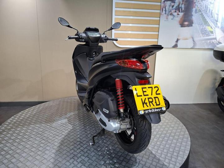 A sleek black scooter displayed from the rear, showcasing its modern design, with a visible license plate reading "LE72 KRD" on a textured circular platform against a neutral background.