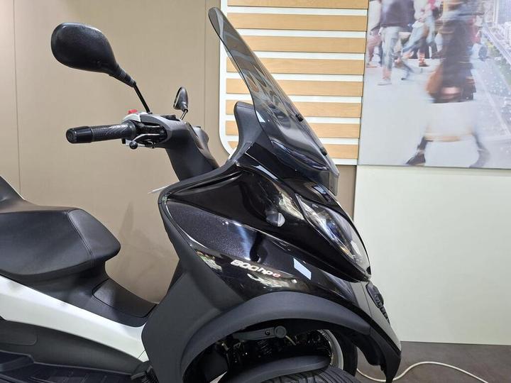 A close-up view of a black scooter with a windshield, showing the handlebar, mirror, and seat, set against a neutral backdrop featuring a blurred image of people walking.