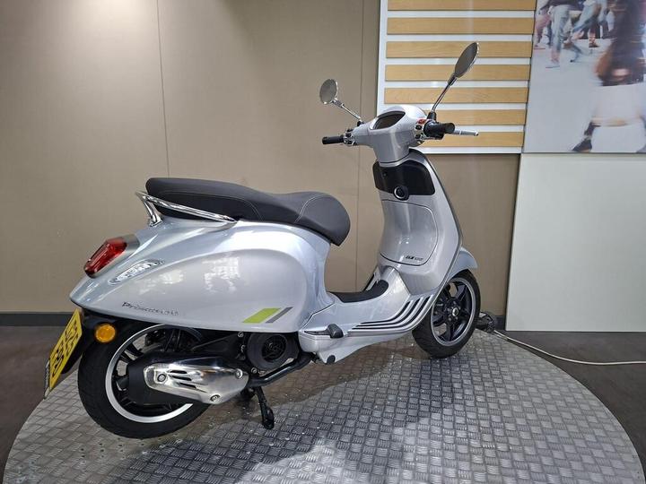 A sleek silver scooter is displayed on a circular metallic stand, showcasing its streamlined design with a black seat, rearview mirrors, and detailed branding on the side. The background features a minimalist design, emphasizing the scooter's modern aesthetics.