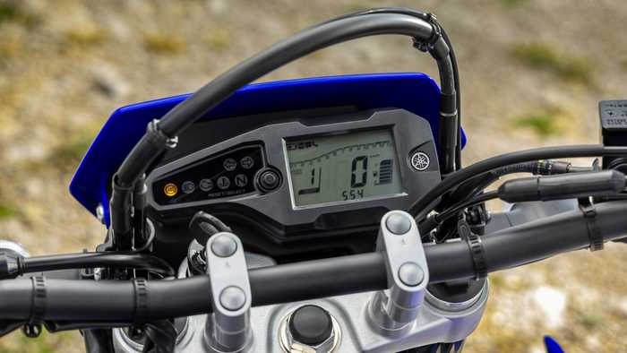 Close-up view of a motorcycle's digital dashboard, displaying information such as speed (0 km/h), gear (1), and odometer (554 km). The dashboard features buttons for resetting and selecting settings, with a blue and black design.