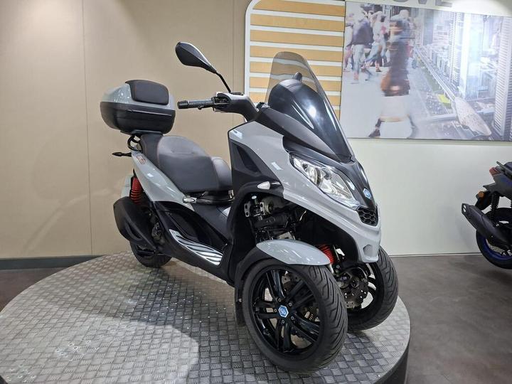 A grey three-wheeled scooter with a sleek design, featuring a large windshield, comfortable seat, and a rear storage compartment, displayed on a metallic circular platform in a showroom.