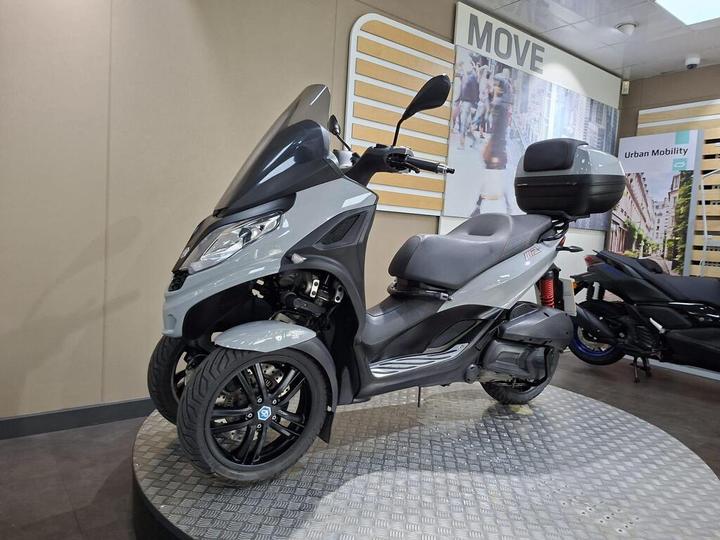 A grey and black three-wheeled scooter is displayed on a circular platform in a modern showroom, featuring a large rear storage compartment and stylish rims, with a background showcasing urban mobility concepts.