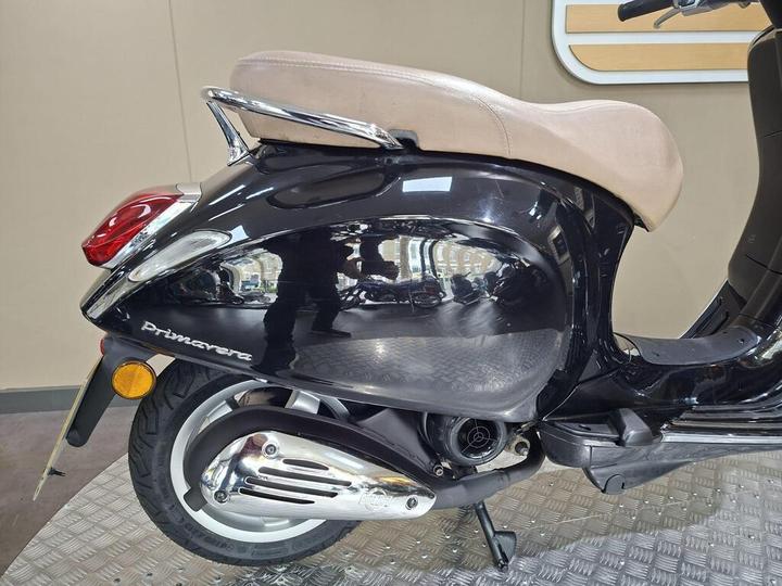 A side view of a black scooter featuring a sleek design, beige seat, and prominent rear tail light, with the word "Primavera" visible on the body. The background is a neutral-colored wall and a textured floor.