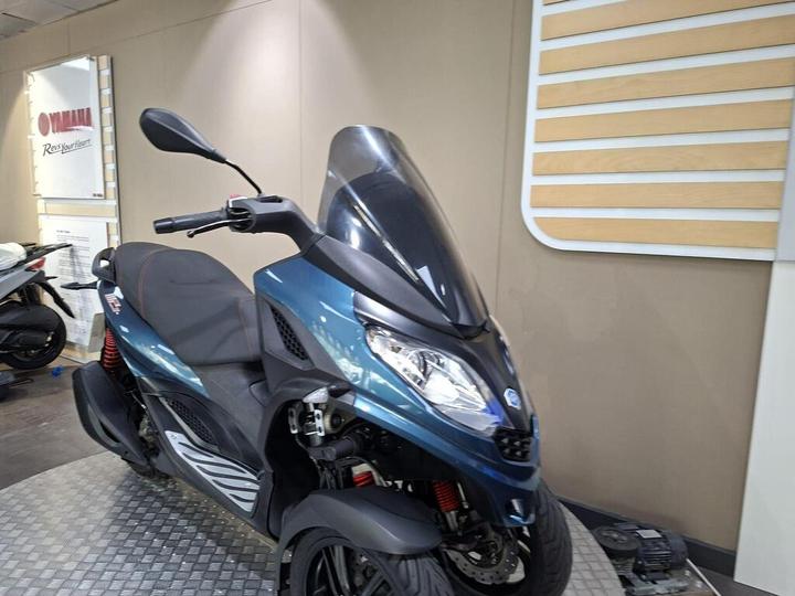 A blue and black scooter is displayed in a showroom, showcasing its sleek design and features, with a shiny front windshield and red shock absorbers. The background includes a Yamaha logo on the wall.