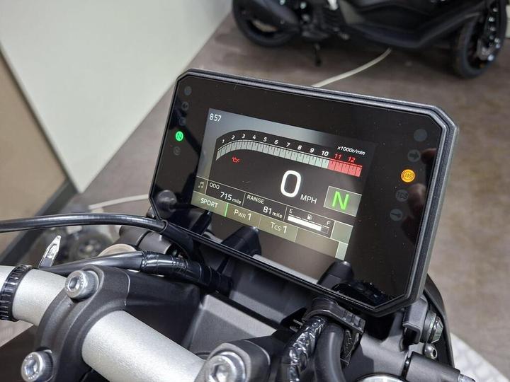 Digital motorcycle display showing speed at 0 MPH, gear indicator N (neutral), and various dashboard readings including odometer at 715 miles, range at 81 miles, and a tachometer scale. The display is mounted on handlebars against a blurred background of a motorcycle.