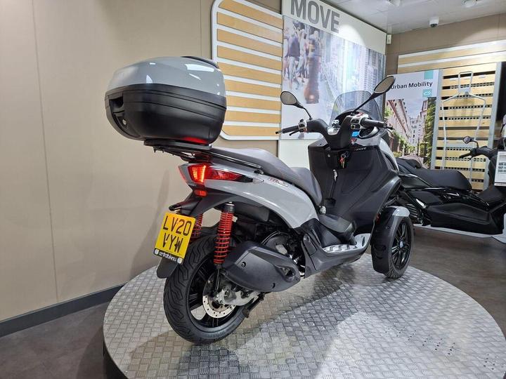 A stylish gray scooter with a rear storage box is displayed in a showroom setting, featuring modern design elements and a vibrant backdrop. The scooter is positioned on a round platform with a textured surface, showcasing its sleek lines and features.