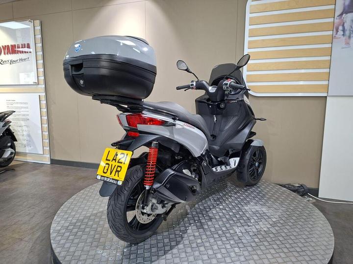 A gray scooter displayed in a showroom, featuring a large rear top case and distinctive design, positioned on a round metallic surface with a textured pattern. The setting includes a Yamaha branding element on the wall.