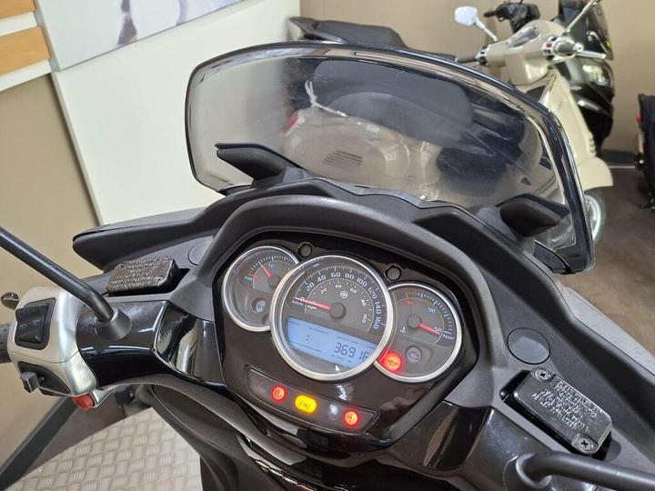 Close-up view of a motorcycle's dashboard, featuring speedometer, fuel gauge, and various indicator lights. The background includes other bikes parked in a garage setting.