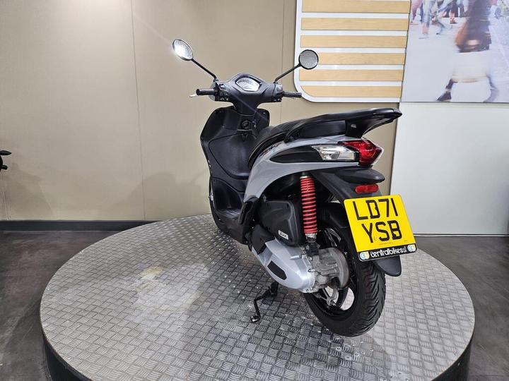 A black and silver scooter displayed on a round, textured platform, viewed from the rear. The scooter features a red shock absorber and has a license plate reading "LD71 YSB." The background includes a neutral wall with wood paneling.