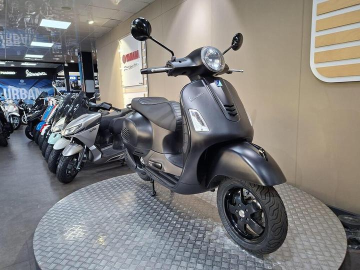 A matte black Vespa scooter displayed on a circular platform in a showroom, surrounded by other scooters in the background.