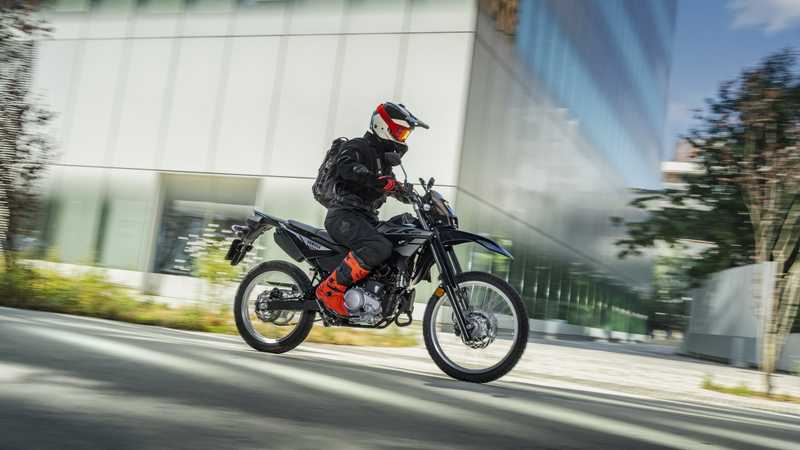 A motorcyclist in black gear and a red helmet rides a black dirt bike on a modern city street, surrounded by glass buildings and greenery, conveying a sense of motion.
